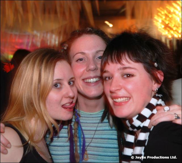 Three smiling young women hug close together, one wearing a black and white striped scarf