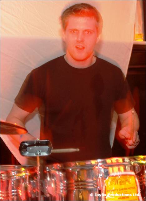 A man in a black t-shirt plays percussion with sticks in front of a white drape