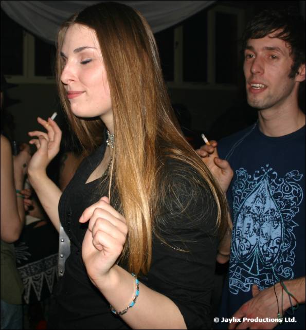 A woman with long hair dances with her eyes closed beside a man holding a cigarette