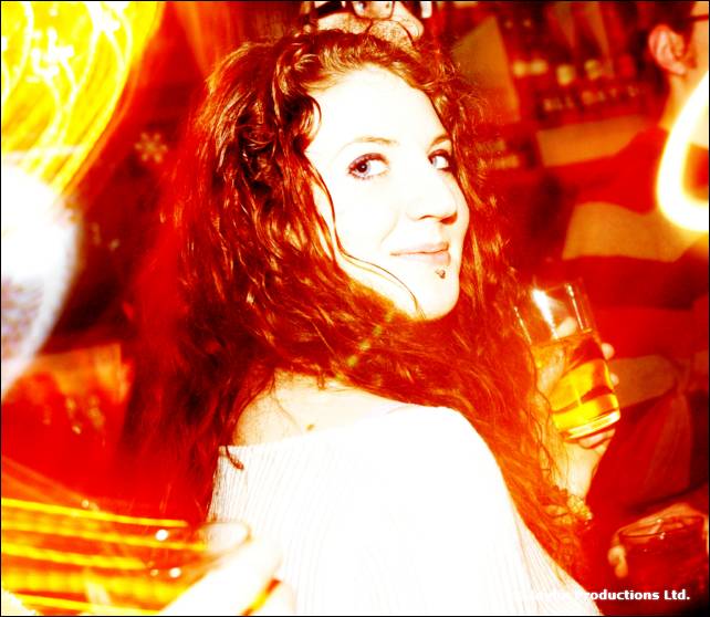 A woman with curly red hair looks over her shoulder holding a drink, in warm orange light