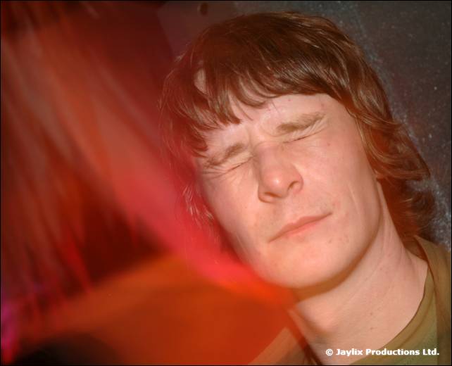 A man with shaggy hair screws his eyes shut, with a red light streak behind him