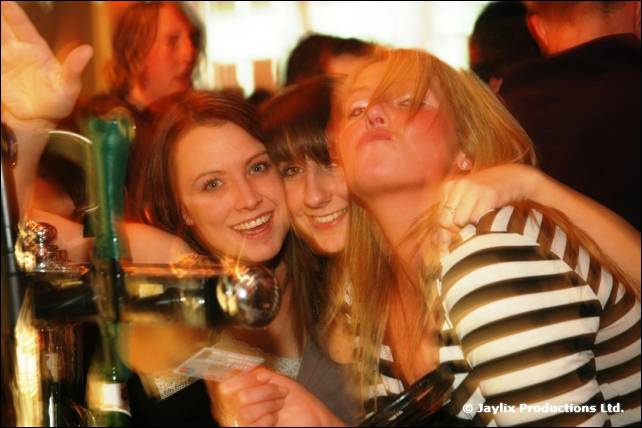 Three young women cheer together behind the bar taps, one in a striped top