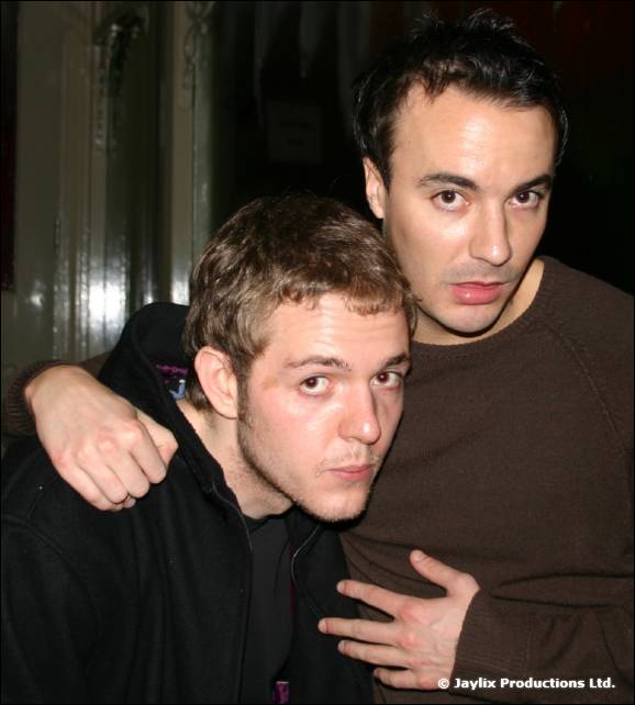 Two men in dark tops stand close with arms around each other, looking unsmiling at the camera