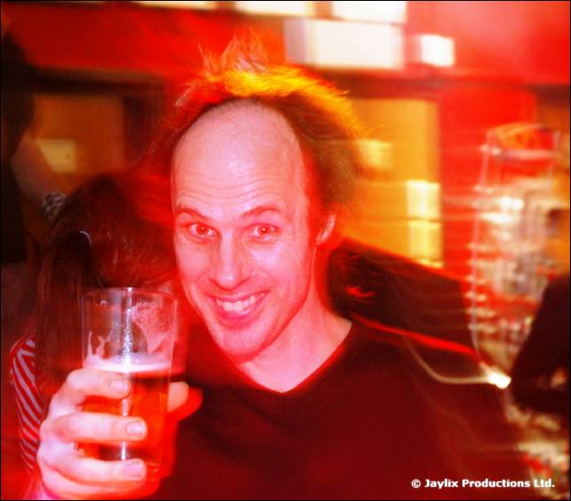 A bald man with spiky hair grins holding a pint of beer, in red light