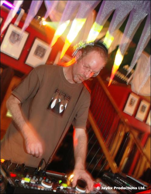 A man in glasses and a brown t-shirt mixes at a DJ desk under hanging streamers
