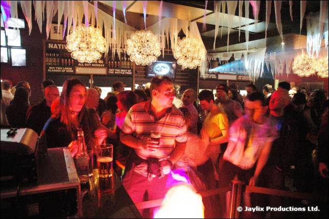 A crowded bar with dancers under chandeliers and white streamers