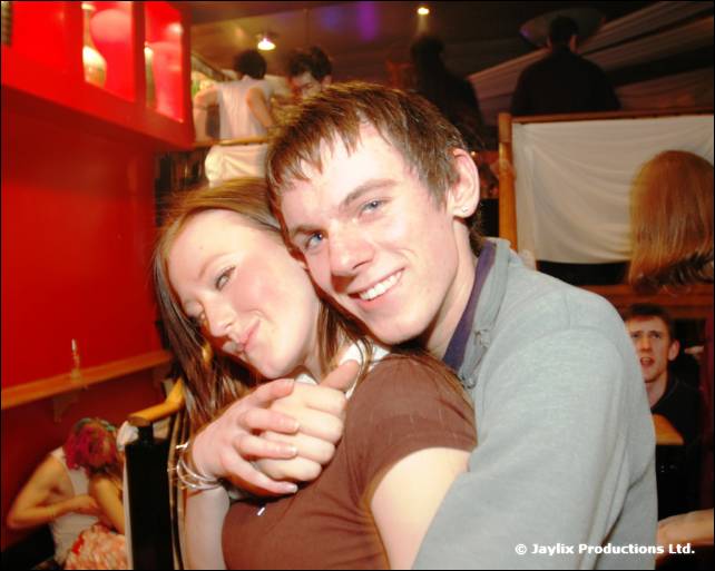 A smiling young man hugs a woman from behind in a red walled bar