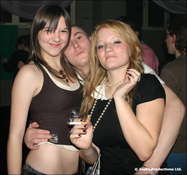 Two young women and a man behind them pose for the camera, one woman holding a cigarette