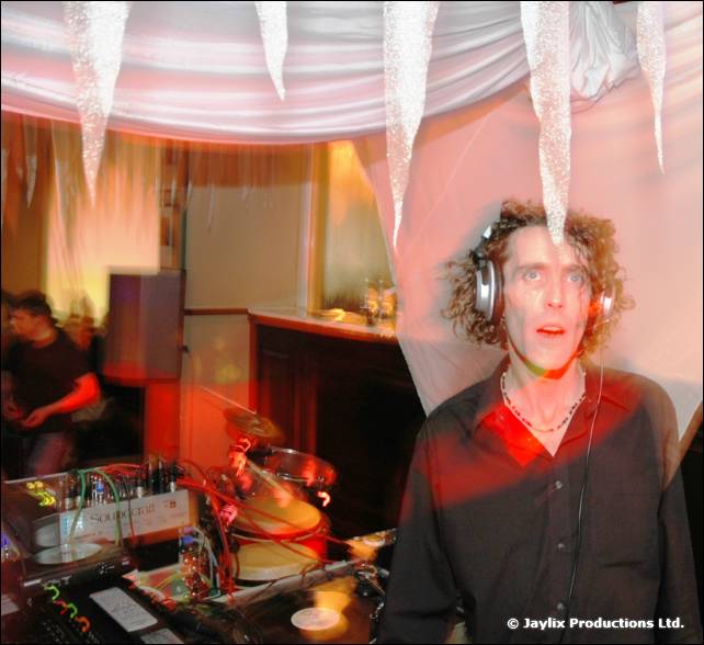 A man with curly hair and headphones stands at DJ decks beneath white hanging streamers