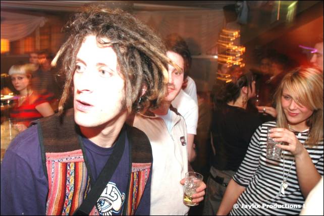 A man with dreadlocks and a patterned waistcoat looks to the side in a busy bar, a woman in a striped top beside him