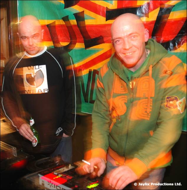 Two bald men at DJ equipment in front of a Bass Culture banner, one in a green hooded top
