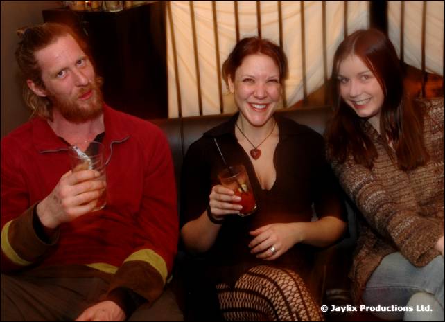 Two women and a bearded man sit on a sofa holding drinks and smiling