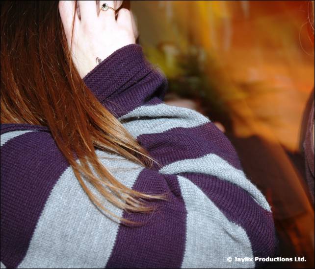 A blurred person with long hair in a purple and grey striped jumper, hand raised to their face