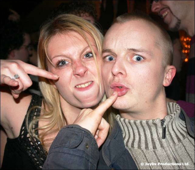 A woman and a man pull faces, each pointing a finger at their own cheek