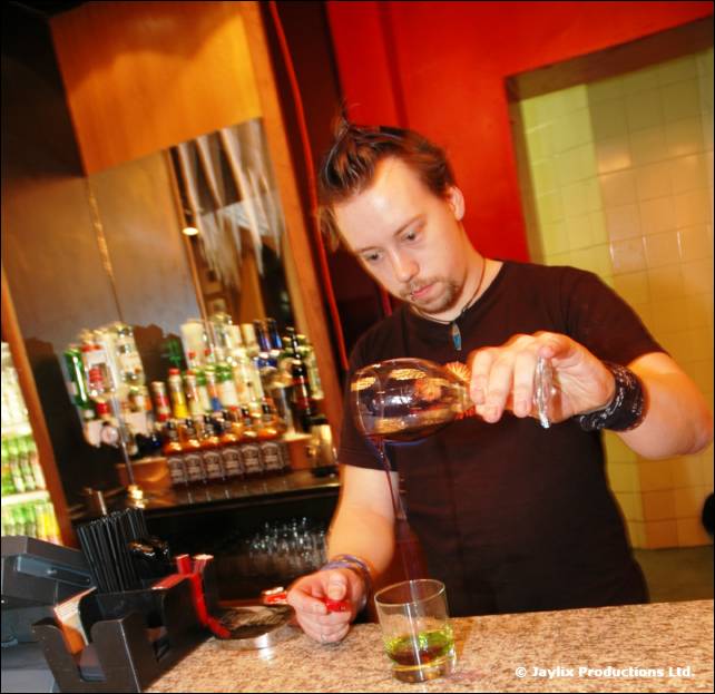 A bartender in a black t-shirt pours a drink from a bottle at a bar