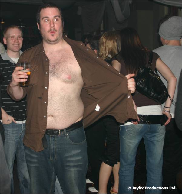 A man in an open brown shirt holds a drink and pulls a surprised face in front of a crowd
