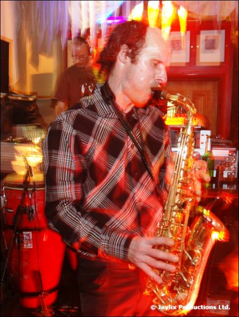 A man in a checked shirt plays a saxophone in front of congas