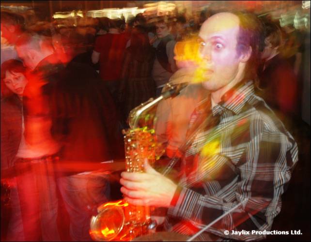 A blurred man in a checked shirt plays a saxophone with a crowd behind
