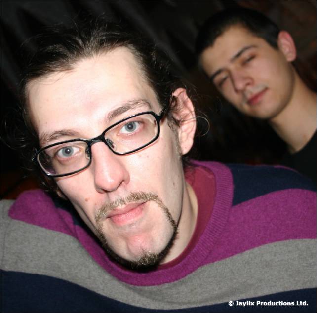 A man with glasses and a goatee looks at the camera, another man with closed eyes behind him