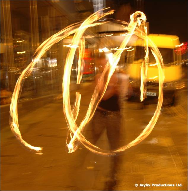 Orange fire trails circle in the air at night on a street, with a dim figure and a van behind