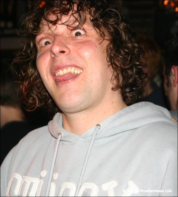 A man with curly hair in a grey hoodie pulls a wide eyed grin at the camera