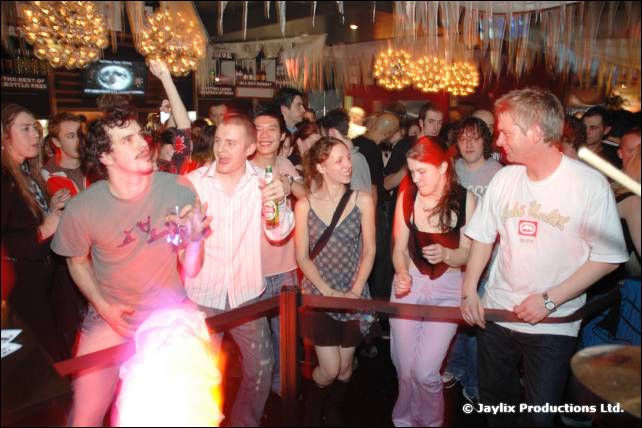 A crowd stands behind a rope barrier under chandeliers, with a bright flame glow at the bottom left