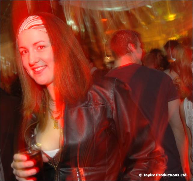 A smiling woman in a patterned headband and black leather jacket holds a drink in a busy room