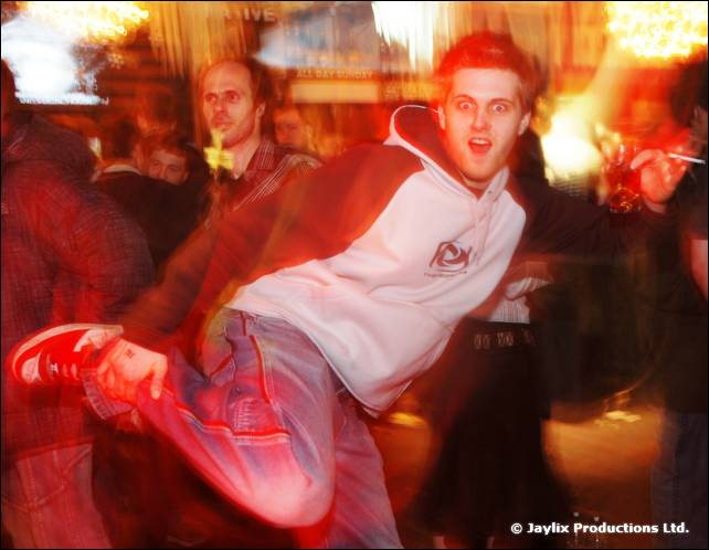 A man in a white and grey hooded top and red trainers kicks a leg out while dancing