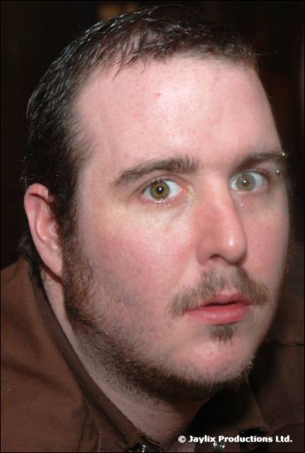 Close-up of a man with short dark hair, stubble and a pierced eyebrow looking at the camera