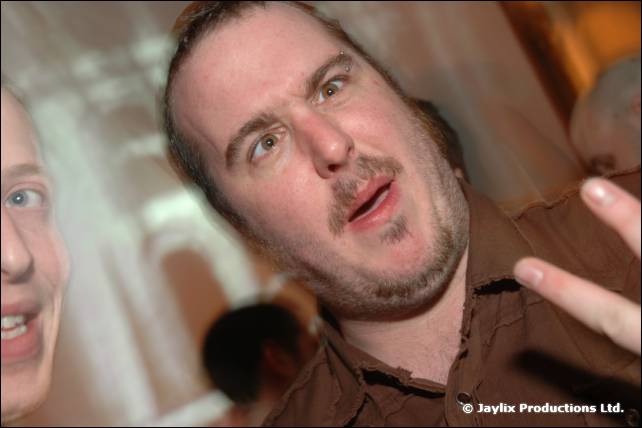 A man with stubble in a brown shirt pulls a face and gestures towards the camera