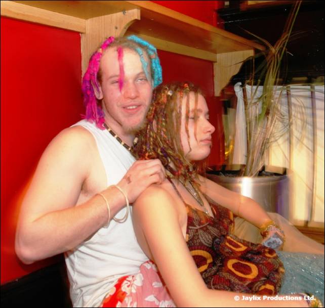 A man with pink and blue dreadlocks in a white vest sits close to a woman with braided hair