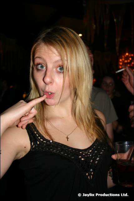 A woman with long blonde hair holds a finger to her lip and looks at the camera
