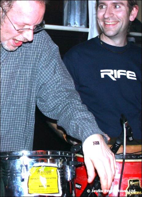 An older man in glasses and a checked shirt plays a conga beside a man in a black RIFE top