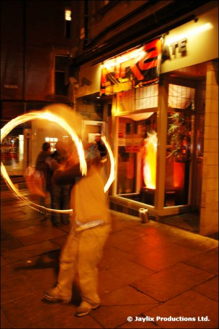 A person spins a fire poi in a circle of light outside a lit shopfront at night