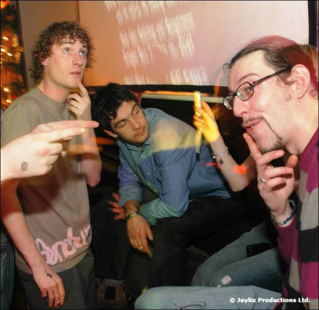 Three men in a dim club, two gesturing with their hands, with a projected screen behind them