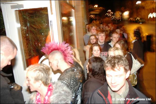 A queue of people waits at a doorway at night, one with a pink mohican