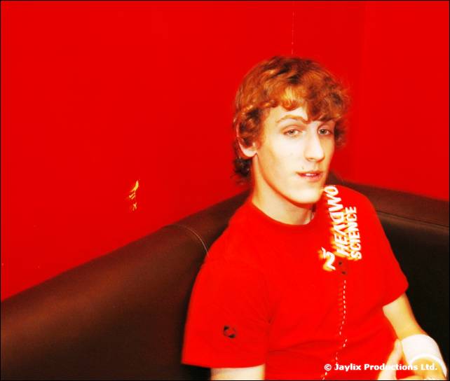 A man with curly ginger hair in a red t-shirt sits on a black sofa against a red wall