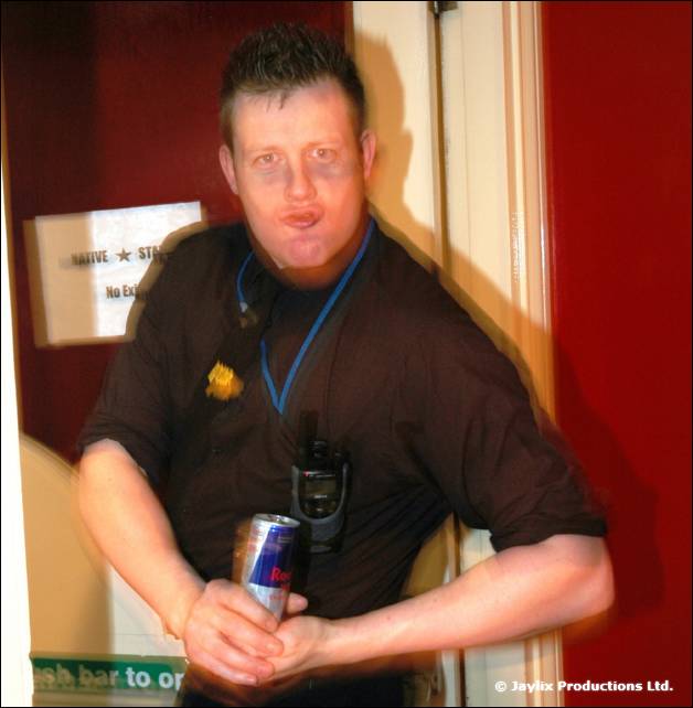 A man in a black shirt with a lanyard holds a can of Red Bull beside a doorway