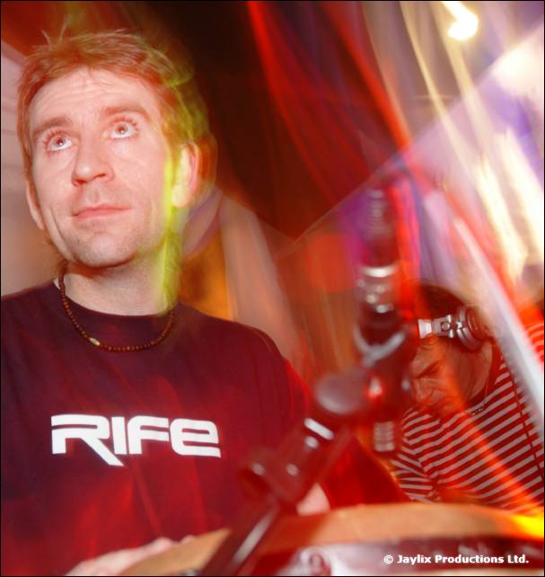 A man in a dark red RIFE t-shirt looks up, with a microphone stand and blurred light streaks