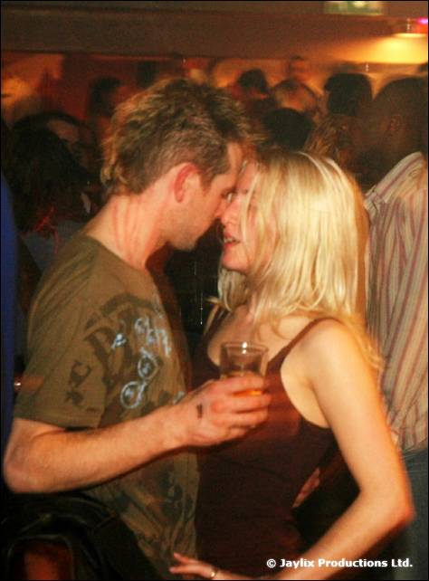 A man and a woman with blonde hair stand face to face holding a drink in a crowd