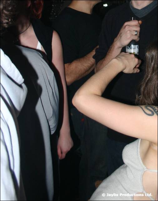 Close view of people standing in a crowd, one holding a bottle and a tattooed arm in the foreground