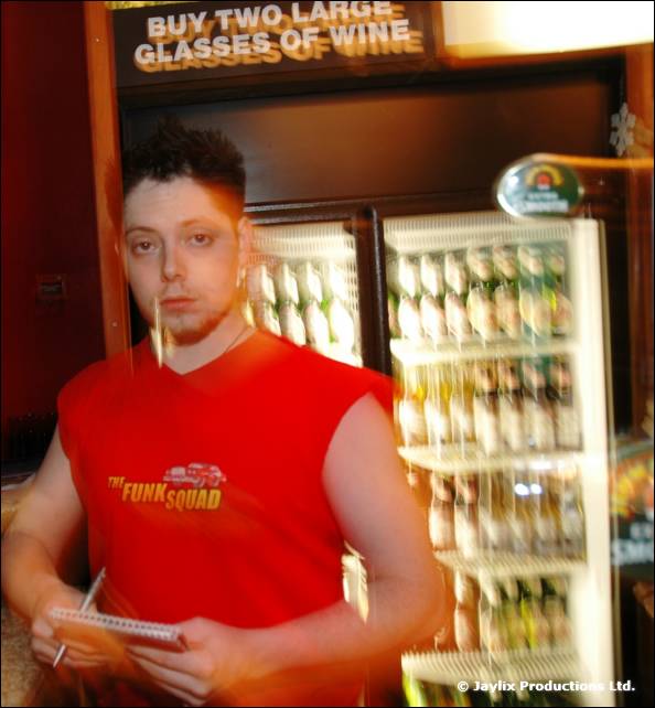 A man in a red sleeveless top holds a notepad in front of drinks fridges under a wine offer sign