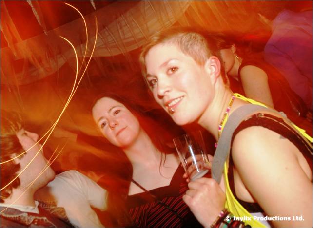 Two people and a person with short hair pose in a dance crowd, with light trails around
