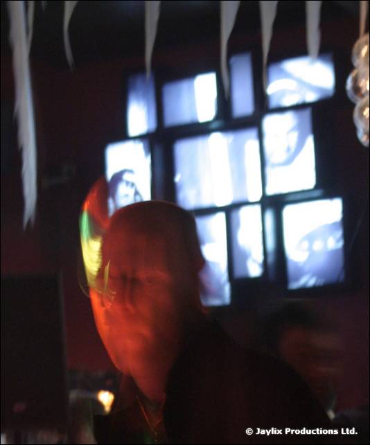 A dark, blurred figure sits in front of a wall of small lit screens