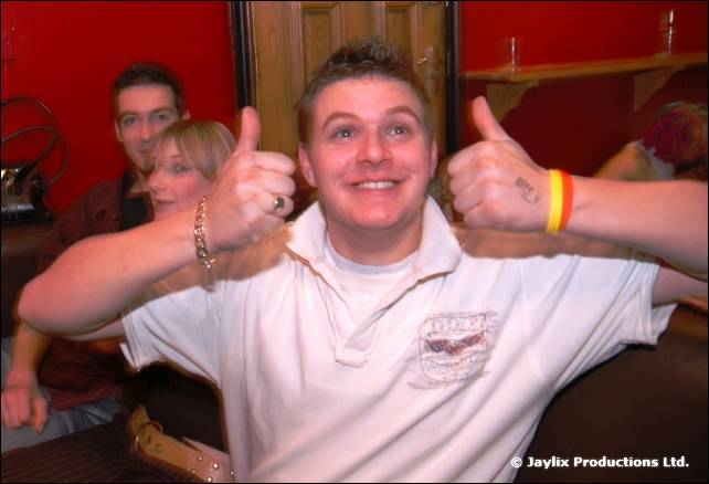 A man in a white polo shirt gives two thumbs up, with others seated behind him in a red walled room