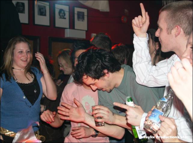 People dance and gesture in a crowded bar, one pointing upwards