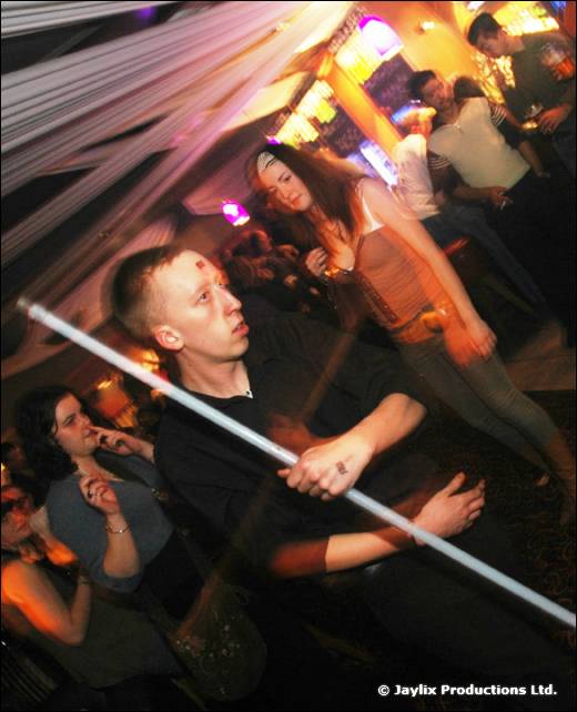 A man in black holds a glowing white staff across his body in a crowd under draped fabric