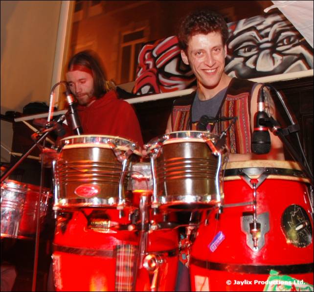 Two men play congas and drums in front of a painted face mural