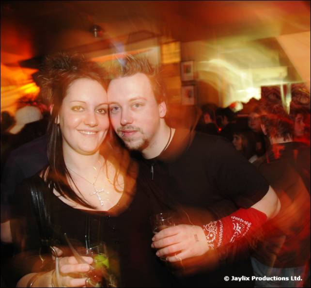 A smiling woman and a man in black with a red bandana on his wrist stand together holding drinks