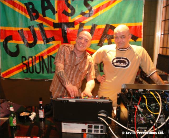 Two bald men stand at DJ equipment in front of a Bass Culture Sound banner